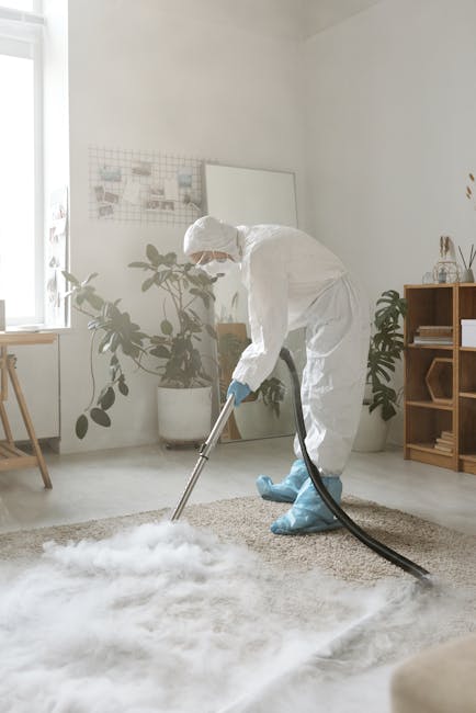 A person dressed in a light-colored jumpsuit is engaged in surface cleaning using a portable vacuum or extractor machine on a dark carpeted floor in an indoor setting, possibly a conference room or of