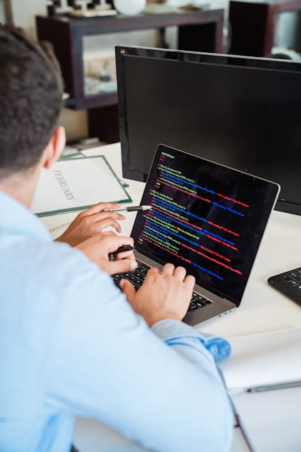 A person working at a desk, viewed from behind, with two monitors and a laptop. The person is using a laptop that displays a screen filled with colorful lines of code. The desk surface is white, and t