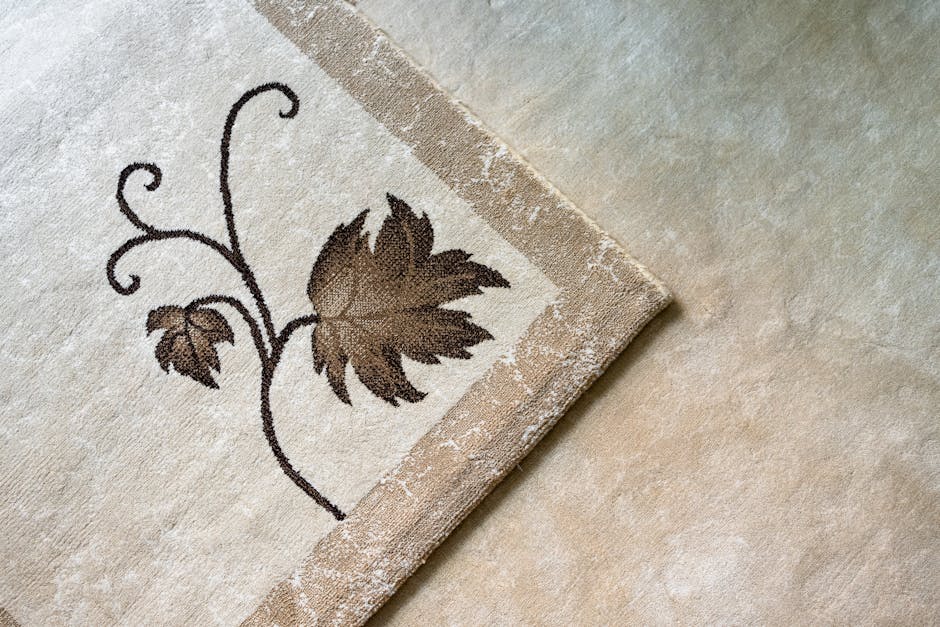 Close-up image of a beige and brown patterned carpet with a floral design, featuring large brown leaves and delicate swirling vine motifs. The carpet has a textured surface with a decorative border al